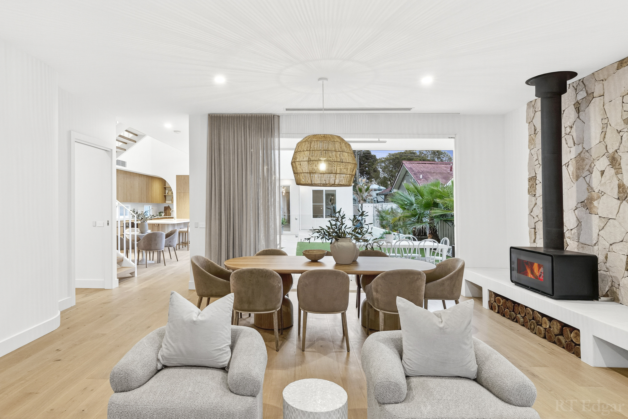Open plan living and dining space with pale timber floors, a stone fireplace and garden outlook